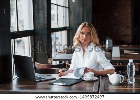 Searching for notes. Businesswoman with curly blonde hair indoors in cafe at daytime.