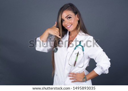 Attractive young doctor woman in casual clothes imitates telephone conversation, keeps hand near ear as if holding mobile phone, has confident facial expression, isolated outdoors. Call me!