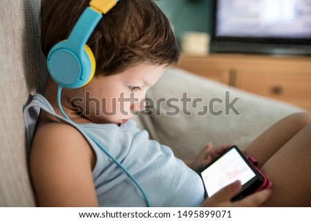 Overstimulated children concept. Too Much Screen Time. 4 years boy in headphones watching videos while tv is working.  Royalty-Free Stock Photo #1495899071