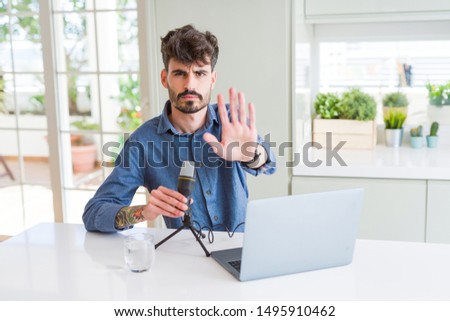 Young man recording podcast using microphone and laptop with open hand doing stop sign with serious and confident expression, defense gesture