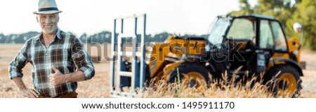 panoramic shot of happy bearded farmer showing thumb up near tractor 
