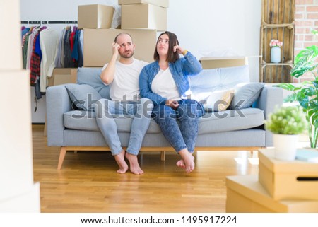Young couple sitting on the sofa arround cardboard boxes moving to a new house confuse and wonder about question. Uncertain with doubt, thinking with hand on head. Pensive concept.