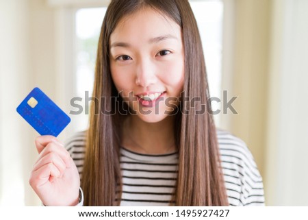 Beautiful Asian woman holding credit card with a happy face standing and smiling with a confident smile showing teeth