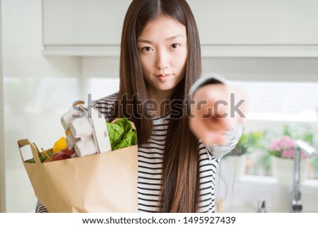 Beautiful Asian woman holding paper bag of fresh groceries pointing with finger to the camera and to you, hand sign, positive and confident gesture from the front