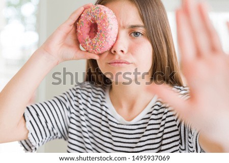 Beautiful young girl kid eating sweet pink donut with open hand doing stop sign with serious and confident expression, defense gesture