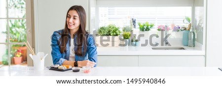 Wide angle picture of beautiful young woman eating delivery sushi looking away to side with smile on face, natural expression. Laughing confident.
