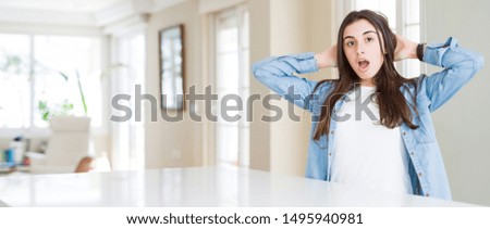 Wide angle picture of beautiful young woman sitting on white table at home Crazy and scared with hands on head, afraid and surprised of shock with open mouth