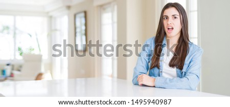 Wide angle picture of beautiful young woman sitting on white table at home In shock face, looking skeptical and sarcastic, surprised with open mouth