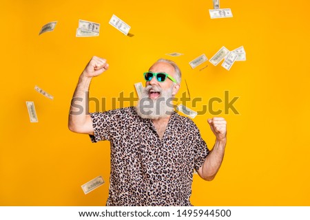 Portrait of crazy retired funky bearded old man with eyeglasses eyewear raise his fists scream yeah celebrate victory look at money falling wearing leopard shirt isolated over yellow background