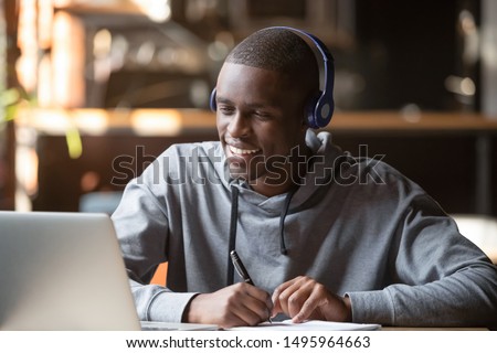 Smiling african young man student wear headphones looking at laptop screen writing notes, happy black guy e-learning in internet study online with teacher on computer watch webinar sit in cafe Royalty-Free Stock Photo #1495964663