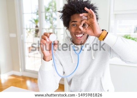 African American man holding stethoscope checking health with happy face smiling doing ok sign with hand on eye looking through fingers