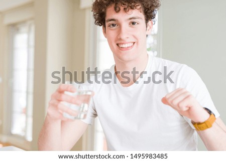 Young man drinking a glass of water at home with surprise face pointing finger to himself