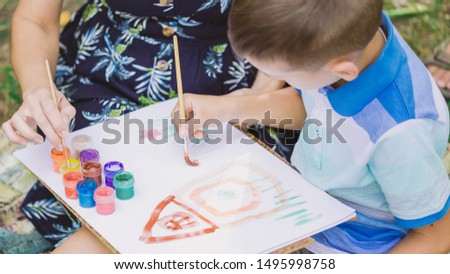 Mother and son painting picture outdoors. Young mom teaching her little son to paint. Baby drawing house and flowers on white sheet of paper. Horizontal color photography.