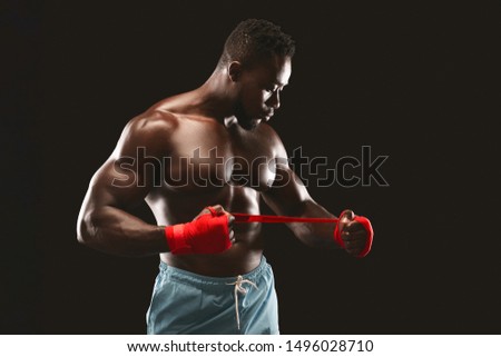 Black young kickboxer getting ready for fight, wrapping his fists, black background, free space
