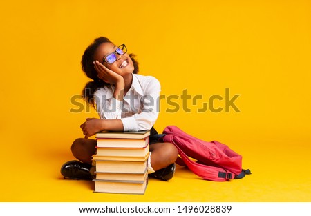 Cheerful African American Schoolgirl Dreaming Sitting At Book Stack Over Yellow Background In Studio. Copy Space Royalty-Free Stock Photo #1496028839