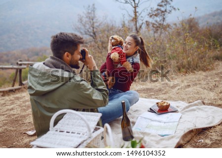 Gorgeous smiling Caucasian brunette sitting on blanket and posing with her dog while her boyfriend taking picture of them. Picnic at autumn concept.