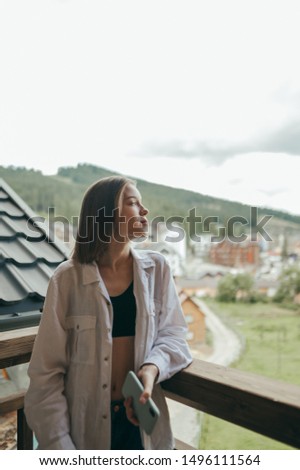 Portrait of attractive girl standing on balcony in hotel in mountains, looking away with serious face. Beautiful woman in stylish clothes relaxing in a countryside in the mountains. Vertical photo.