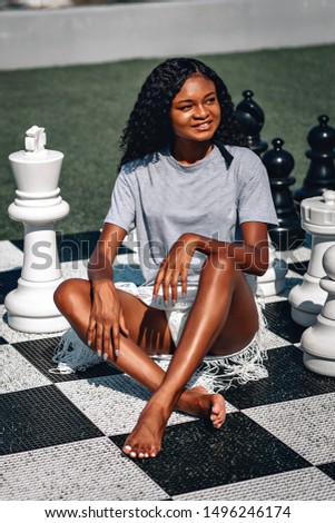 Smart African-American woman playing giant chess as she sits on the board with her legs crossed; glamourous intelligence concept.
