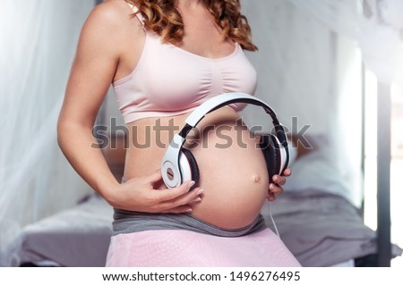 Pregnant woman feeling happy at home while taking care of her child. Pregnant woman holding headphones on her belly. Maternity prenatal care and woman pregnancy concept.