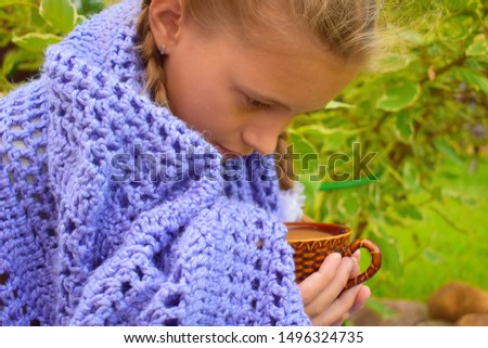 Beautiful girl in a knitted blanket drinks hot tea. Cold evening with a cup of coffee. A cozy picture with a cute girl with pigtails