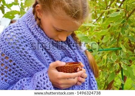Beautiful girl in a knitted blanket drinks hot tea. Cold evening with a cup of coffee. A cozy picture with a cute girl with pigtails