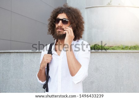 Outdoor portrait of handsome good looking man with lush beard and curls talking on phone while walking down the street, wearing casual clothes