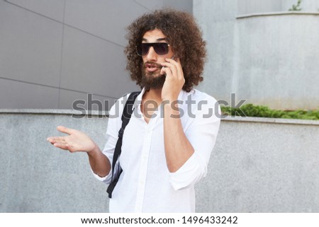 Young handsome bearded guy with curly hair posing over city background with agitated face, wrinkling forehead and gesticulating while explaining something on phone