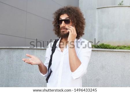 Wonderstrucked young bearded man standing over outdoor backgroung, raising his palm and wrinkling forehead, wearing glasses and white shirt