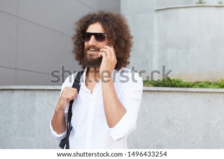 Happy young bearded male walking down the street and talking on phone, wearing sunglasses and casual clothes, being cheerful and pleased