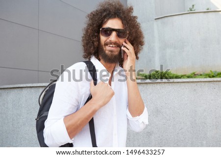 Attractive curly young male with lush beard walking down the street on warm day, going to make call with his mobile phone, being in nice mood and smiling broadly to camera