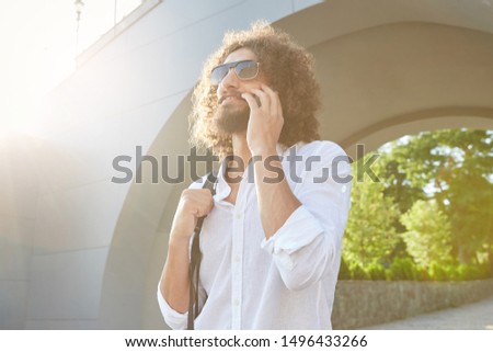 Outdoor shot of handsome pretty bearded male with curls talking on mobile phone, walking through city park, wearing sunglasses and casual clothes