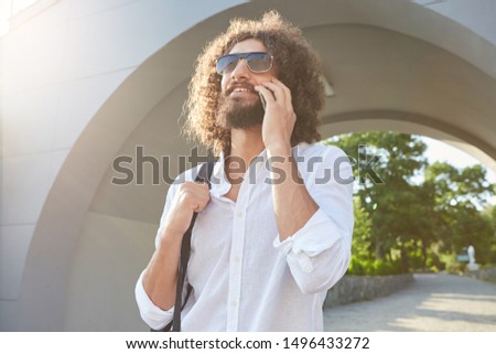 Happy lovely bearded man with curly hair wearing sunglasses, walking in city park on sunny day while talking on mobile phone, holding his backpack