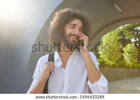 Young attractive bearded man with brown curly hair having pleasant conversation on phone, smiling cheerfully, posing over city park on sunny day