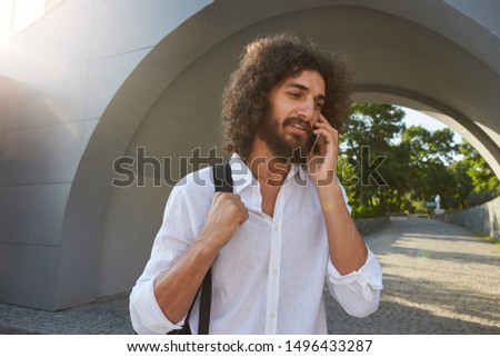 Young beautiful male with lush beard and brown curly hair posing over arch in green park on sunny warm day, making call with his mobile phone