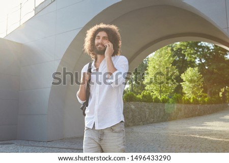 Indoor photo of beautiful young bearded male with brown curly hair walking in green city park on sunny warm day, looking cheerfully to camera while making call with his mobile phone