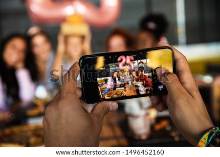 Close up of hands with phone taking picture of group of young friends having fun in restaurant, talking and laughing while dining at table.