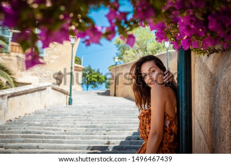 young brunette woman wearing a floral pattern summer dress, leaning against a lamp post next to a stone wall and colorful blooming bushes, in Artà on Mallorca