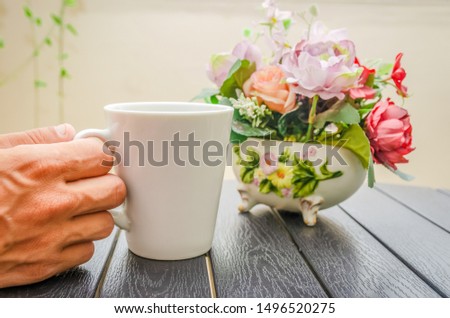 A man's hand holds a white Cup of coffee on a wooden table close-up, next to a bouquet of flowers