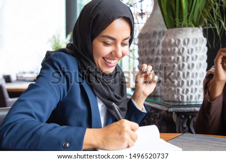 Happy businesswoman taking notes in coffee shop. Young Muslim business woman in hijab and office suit writing in notebook and laughing. Muslim business professional concept