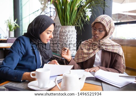 Female business colleagues analyzing and discussing documents in cafe. Muslim businesswomen sitting in coffee shop, working on papers and talking. Paperwork concept