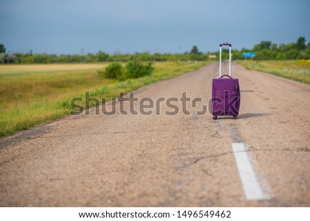 Purple suitcase on wheels with a raised handle stands in the middle of an empty asphalt road in the fields. Summer, Sunny weather, travel.