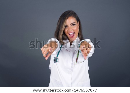 Close-up portrait of surprised pretty young doctor woman in casual clothes pointing with two fingers to the camera saying: I choose you!, looking up with open mouth, isolated over gray background