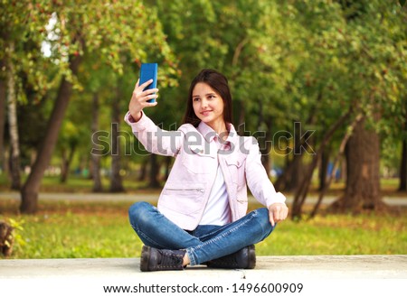 Young beautiful girl photographs herself on a cell phone while sitting in an autumn park.