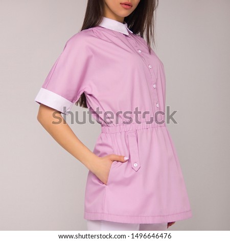 close-up young stylish nurse with long brunette hair in pink medical costume is standing with hand in her pocket on white wall background. medical fashion concept. free space