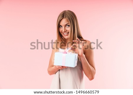 Image of alluring young woman wearing dress expressing surprise and holding gift box isolated over pink background