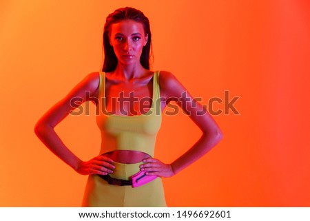 Photo of attractive brunette woman wearing formfitting sportswear standing with arms on waist isolated over orange neon wall