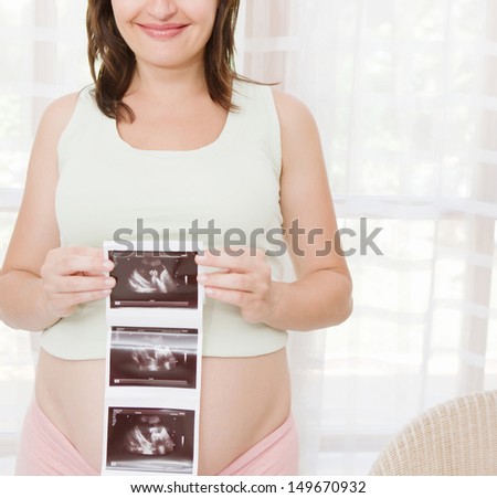 Portrait of a pregnant woman proudly holding the echography digital photograph scans of her unborn baby in front of her belly at home, smiling.