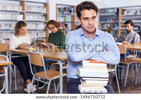 Portrait of weary upset man with stack of books in hands in public library