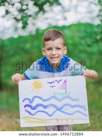 Closeup view of happy smiling and laughing emotional white little kid of 5 year old looks at camera holding big sheet of paper with cute painted picture of small sailing boat, sea water, beach and sun
