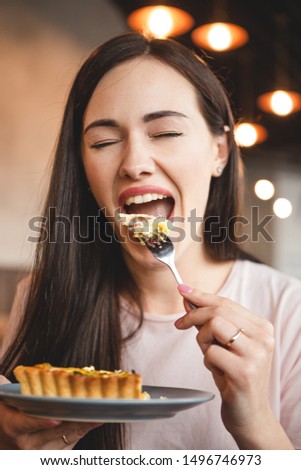 woman with exquisite smile and beautiful eyes having breakfast with her friend in a cozy cafe. She eats a fork a piece of cake and drinks tea sitting in a chair at a table